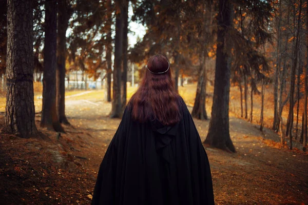Jovem mulher bonita e misteriosa na floresta, em manto preto com capuz, imagem de elfo da floresta ou bruxa, de volta — Fotografia de Stock