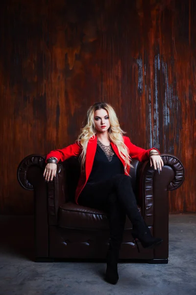 Mujer rubia joven y atractiva en chaqueta roja se sienta en sillón de cuero, fondo grunge pared oxidada —  Fotos de Stock