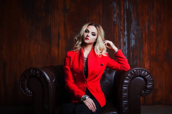 Mladá a atraktivní blond žena v červené bundě sedí v koženém křesle, pozadí grunge rezavé zdi — Stock fotografie
