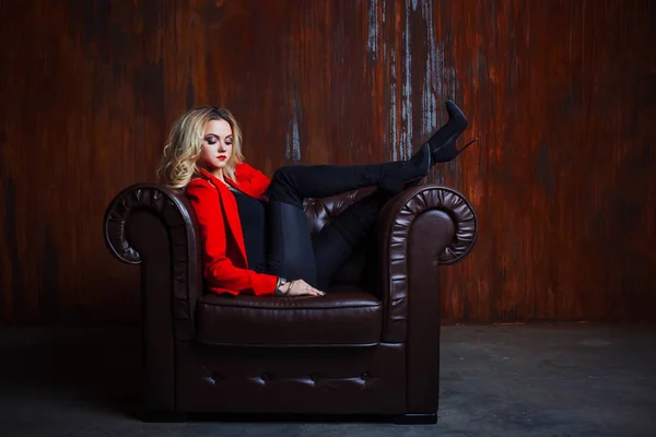 Młody i atrakcyjny blond kobieta w czerwonej kurtce siedzi w skórzany fotel, stopy na podłokietniku — Zdjęcie stockowe