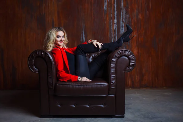 Jeune et jolie femme blonde en veste rouge assise dans un fauteuil en cuir, pieds sur l'accoudoir — Photo