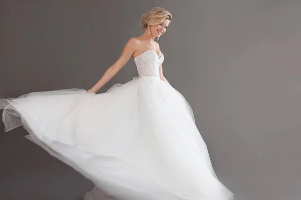 Танцующая молодая невеста в роскошном свадебном платье. Красивая девушка в белом. Эмоции счастья, смеха и улыбки, серый фон — стоковое фото