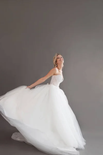 Dansende jonge bruid in luxe trouwjurk. Mooi meisje in het wit. Emoties van geluk, gelach en glimlach, grijze achtergrond — Stockfoto