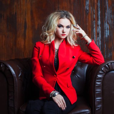 Genç ve çekici sarışın kadın kırmızı ceketli oturur deri koltuk, arka plan grunge paslı duvar