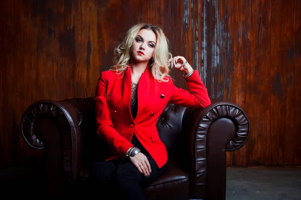 Ung och attraktiv blond kvinna i röd jacka sitter i läderfåtölj, grunge rostig fondvägg — Stockfoto