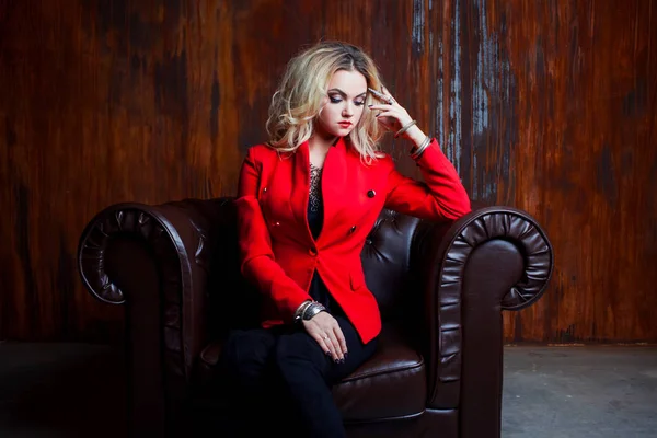 Young and attractive blond woman in red jacket sits in leather armchair, background grunge rusty wall — Stock Photo, Image