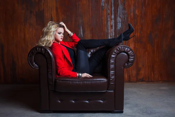 Young and attractive blond woman in red jacket sits in leather armchair, feet on the armrest — Stock Photo, Image