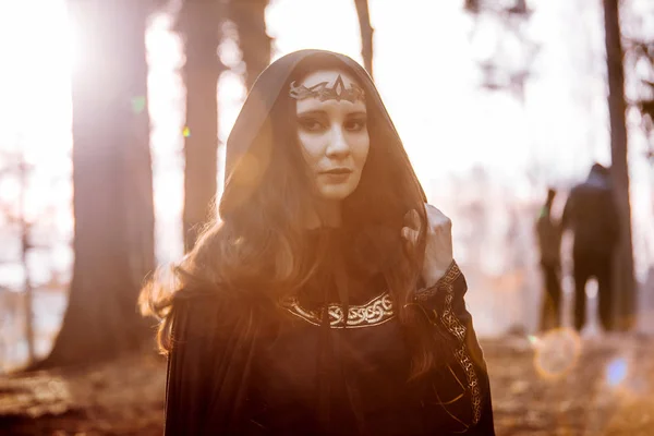 Молодая красивая и таинственная женщина в лесу, в черном плаще с капюшоном, образ лесного эльфа или ведьмы — стоковое фото