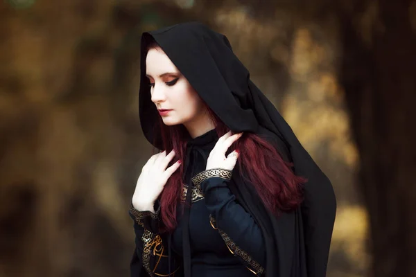 Jovem mulher bonita e misteriosa na floresta, em manto preto com capuz, imagem de elfo da floresta ou bruxa — Fotografia de Stock