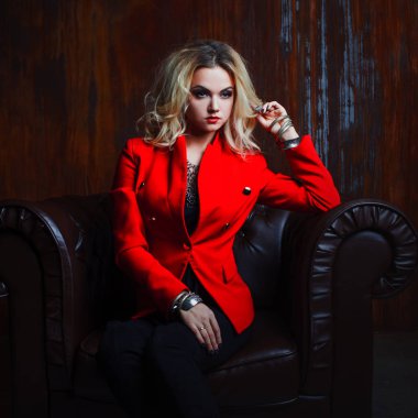 Genç ve çekici sarışın kadın kırmızı ceketli oturur deri koltuk, arka plan grunge paslı duvar