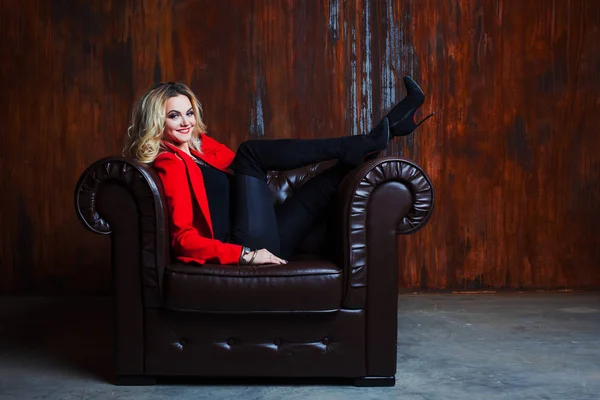 Молода і приваблива блондинка в червоній куртці сидить у шкіряному кріслі, ноги на підлокітнику — стокове фото