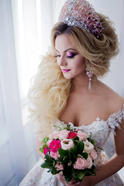 Jonge bruid met perfecte huid mode en make-up, krullend haar, bloemen en tiara op het hoofd, tegen een raam binnenshuis — Stockfoto