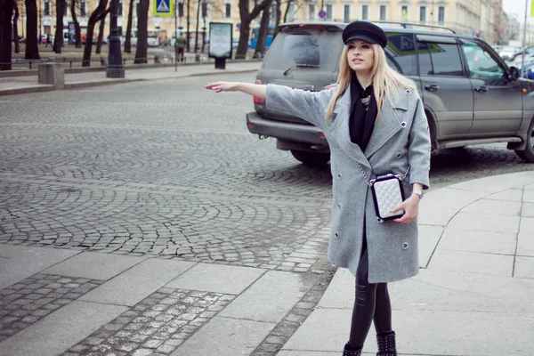 Jovem, quadril e atraente táxi saudação loira, menina em um chapéu elegante e um casaco cinza — Fotografia de Stock
