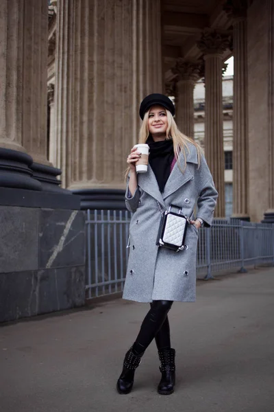 Молода, хіп-хоп і приваблива блондинка, що гуляє по місту з кавою, щоб піти, дівчина в стильному капелюсі і сіре пальто — стокове фото