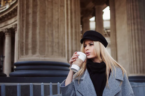 Młodych, hip i atrakcyjna blondynka chodząc po mieście z kawy iść, Dziewczyna w stylowy kapelusz i płaszcz szary — Zdjęcie stockowe