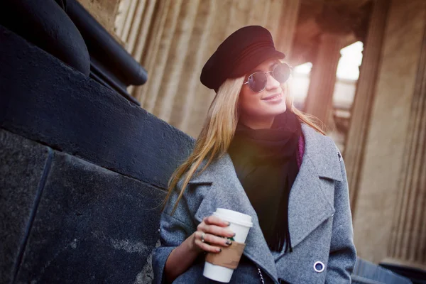 Junge, hippe und attraktive Blondine, die mit Coffee to go durch die Stadt läuft, Mädchen mit stylischem Hut und grauem Mantel — Stockfoto