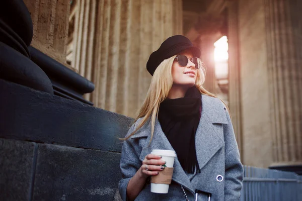 Junge, hippe und attraktive Blondine, die mit Coffee to go durch die Stadt läuft, Mädchen mit stylischem Hut und grauem Mantel — Stockfoto