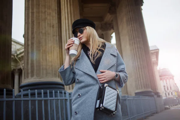 Joven, cadera y atractiva rubia paseando por la ciudad con café para llevar, chica con un sombrero elegante y un abrigo gris — Foto de Stock