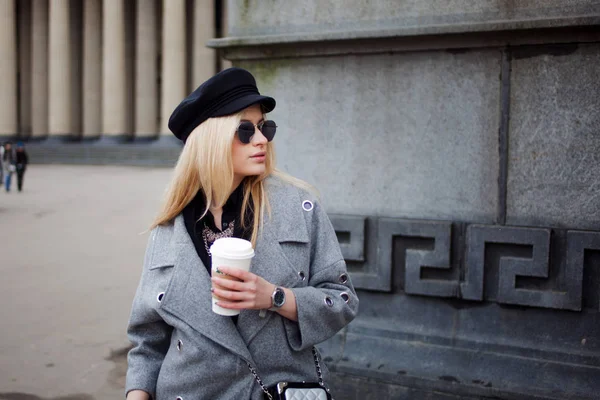 Junge, hippe und attraktive Blondine, die mit Coffee to go durch die Stadt läuft, Mädchen mit stylischem Hut und grauem Mantel — Stockfoto