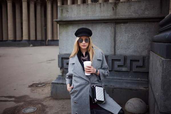 Junge, hippe und attraktive Blondine, die mit Coffee to go durch die Stadt läuft, Mädchen mit stylischem Hut und grauem Mantel — Stockfoto