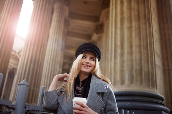 年轻、 髋关节和吸引力金发在城市与咖啡走来走去，在一顶时髦的帽子和一件灰色的大衣的女孩 — 图库照片