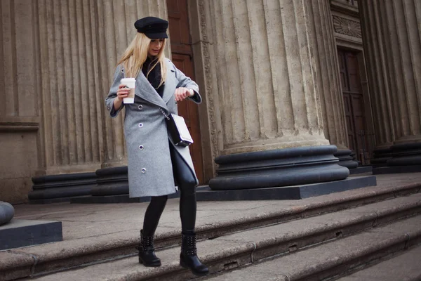 Молодая, модная и привлекательная блондинка гуляет по городу с кофе на вынос, смотрит на часы и спешит по делам — стоковое фото