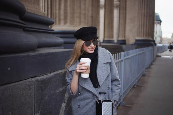 若い、ヒップで魅力的な金髪のコーヒーを飲みながら街を歩いて行き、スタイリッシュな帽子および灰色のコートの女の子 — ストック写真