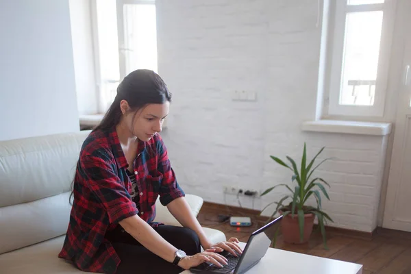 Joven chica hipster en un sofá blanco con portátil. La mujer freelancer trabaja en casa. Joven morena atractiva en la camisa a cuadros rojo en el interior moderno en el sofá blanco. Tono suave y enfoque —  Fotos de Stock