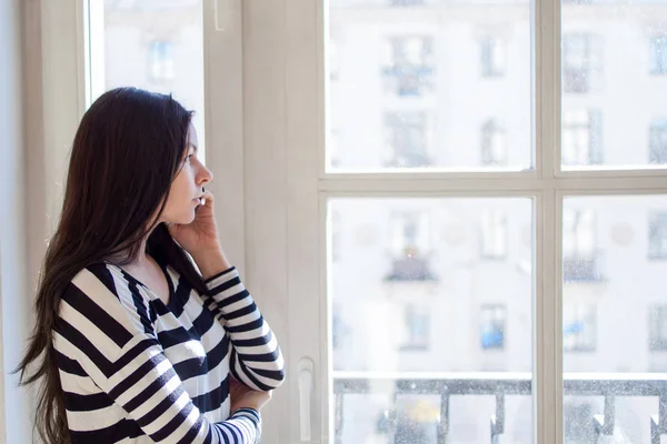 Meisje bij het venster praten aan de telefoon thuis in de ochtendzon. Profile, serieus kijkt naar de afstand — Stockfoto