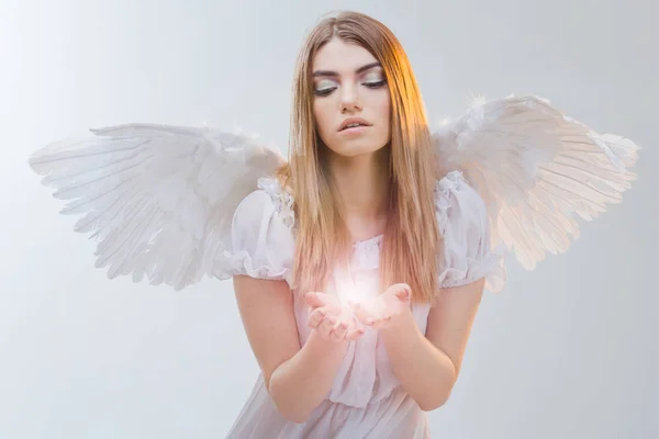 Een engel uit de hemel die houden van licht op de handpalmen. Jonge, prachtige blonde meisje in de afbeelding van een engel met witte vleugels. — Stockfoto