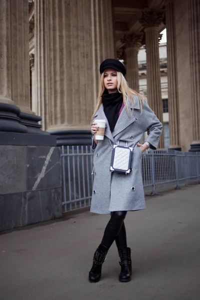 Молодая, модная и привлекательная блондинка, гуляющая по городу с кофе на вынос, девушка в стильной шляпе и сером пальто — стоковое фото