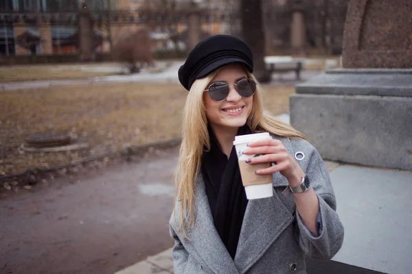 Młodych, hip i atrakcyjna blondynka chodząc po mieście z kawy iść, Dziewczyna w stylowy kapelusz i płaszcz szary — Zdjęcie stockowe