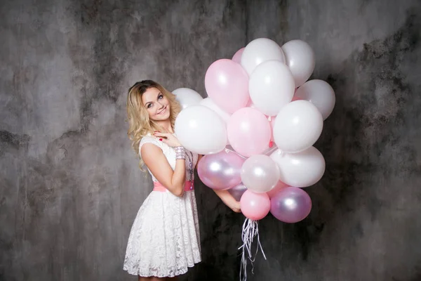 Charmante jonge blonde in een witte jurk met roze sjerp, een grote bundel van ballonnen op het feest te houden. — Stockfoto