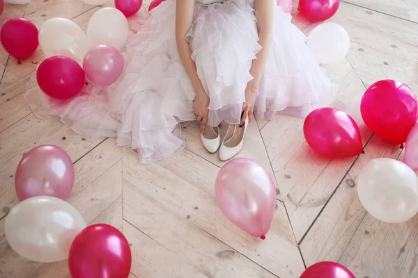 Young woman in wedding dress in luxury interior with a mass of pink and white balloons, sitting on the floor. Hold in hands her white shoes. — Stock Photo, Image