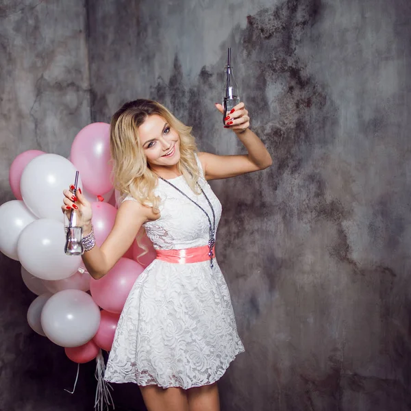 Charmante jonge blonde in een witte jurk met roze sjerp, dansen met twee flessen frisdrank in haar handen Happy en vrolijk meisje met ballonnen. Op grijze gestructureerde achtergrond — Stockfoto