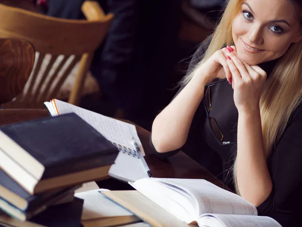 Young woman student of the University. Preparing exam and learning lessons in public library. Sitting at table surrounded by large number of textbooks — Stock Photo, Image