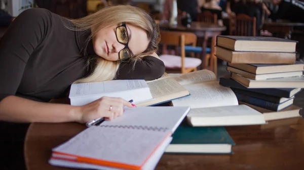 Trött ung kvinna student vid universitetet. Förbereda tentamen och lärdomar i offentligt bibliotek. Sitter vid bord omgiven av stort antal läroböcker — Stockfoto