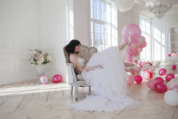 Mujer joven en vestido de novia en el interior de lujo con una masa de globos rosados y blancos, sentado en la silla. Encantadora joven novia morena con corte de pelo corto en vestidos de Quinceañera con estilo . —  Fotos de Stock