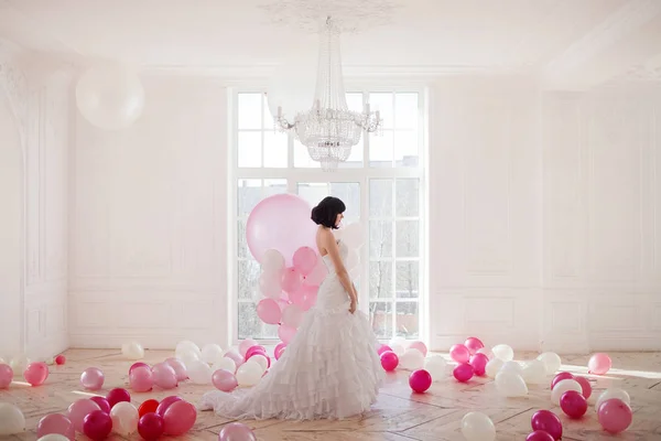 Giovane donna in abito da sposa in interni di lusso con una massa di palloncini rosa e bianchi, in piedi contro la finestra . — Foto Stock