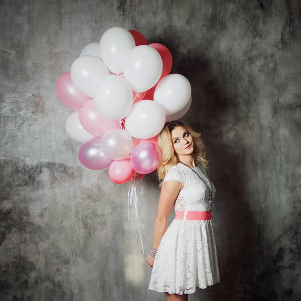 Charmante jonge blonde in een witte jurk met roze sjerp, een grote bundel van ballonnen op het feest te houden. — Stockfoto