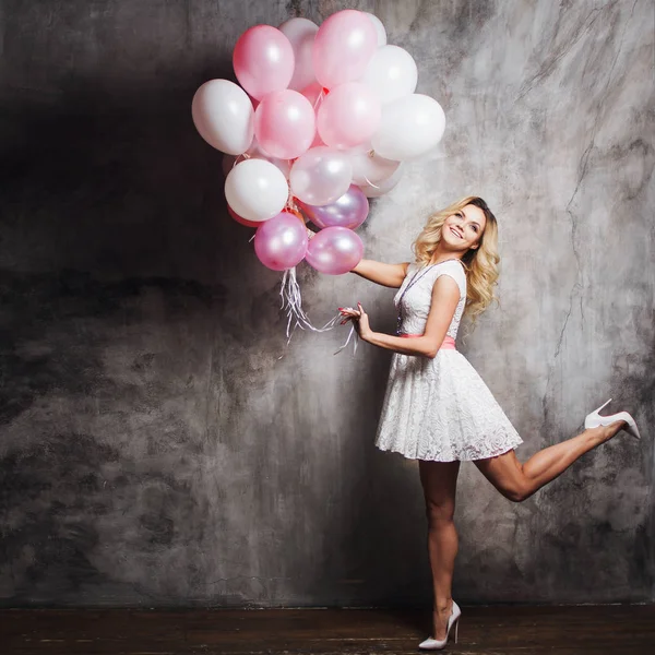 Charmante jonge blonde in een witte jurk met roze sjerp, een grote bundel van ballonnen op het feest te houden. — Stockfoto