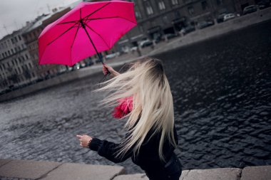 Güzel genç ve mutlu sarışın kadın bir parlak pembe eşarp, lastik çizmeler ve yağmurlu bir şehirde yürüyüş şemsiye.