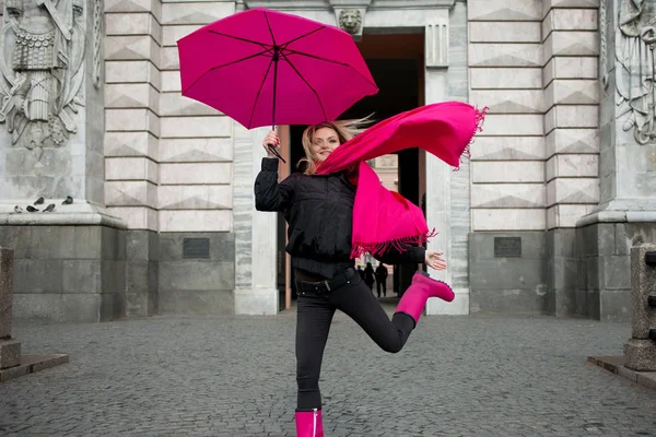 Schöne junge und glückliche blonde Frau mit buntem Regenschirm auf der Straße. Mädchen in leuchtend rosa Schal, Gummistiefeln und Regenschirm laufen in einer verregneten Stadt. — Stockfoto