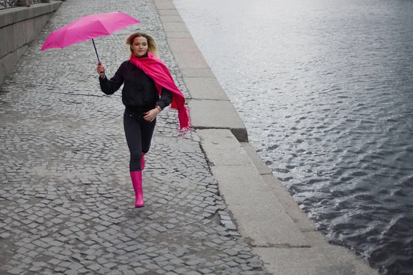 Mulher loira jovem e feliz bonita com guarda-chuva colorido na rua. Menina em um lenço rosa brilhante, botas de borracha e guarda-chuva andando em uma cidade chuvosa . — Fotografia de Stock