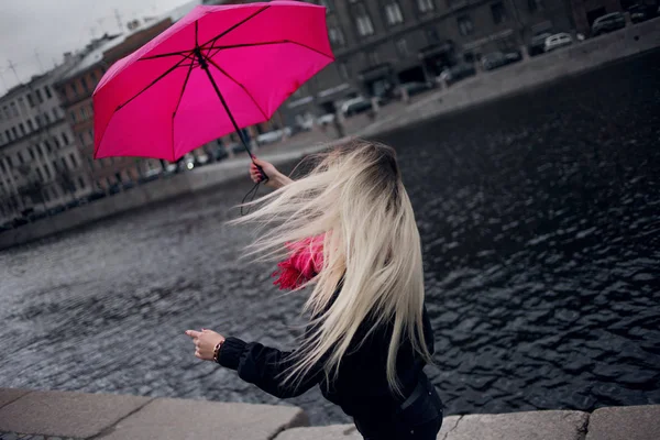 Красивая молодая и счастливая блондинка в ярко-розовом шарфе, резиновых сапогах и зонтике, гуляющая по дождливому городу . — стоковое фото