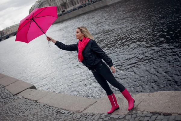Güzel genç ve mutlu sarışın kadın bir parlak pembe eşarp, lastik çizmeler ve yağmurlu bir şehirde yürüyüş şemsiye. — Stok fotoğraf