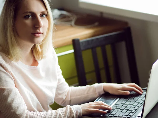 Mujer rubia joven utiliza un ordenador portátil, sentado en casa. Concepto de trabajo en Internet — Foto de Stock