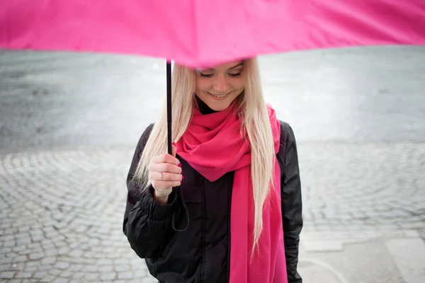 Triste fille chère debout sous un parapluie lumineux dans la rue. Le concept de positivité et d'optimisme. Fille dans une écharpe rose vif et parapluie marchant dans une ville pluvieuse . — Photo