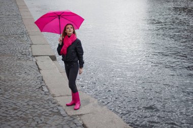 Güzel genç ve mutlu sarışın kadın bir parlak pembe eşarp, lastik çizmeler ve yağmurlu bir şehirde yürüyüş şemsiye.