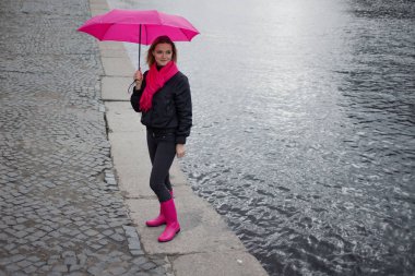 Güzel genç ve mutlu sarışın kadın bir parlak pembe eşarp, lastik çizmeler ve yağmurlu bir şehirde yürüyüş şemsiye.